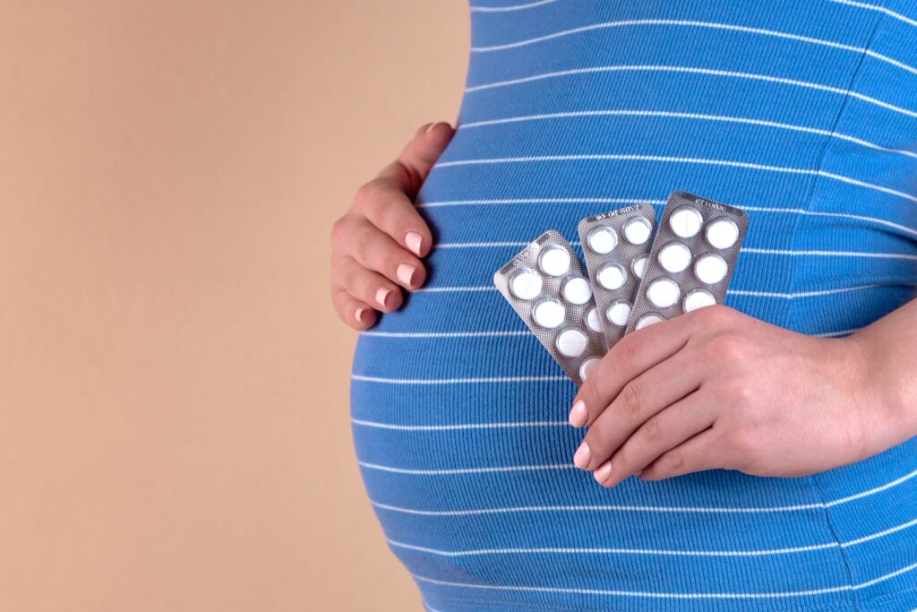 A close-up view of the belly of a pregnant woman in a blue T-shirt, which holds pills in her hands. The concept of medicine during pregnancy
